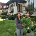 A woman standing in front of a house with bushes.